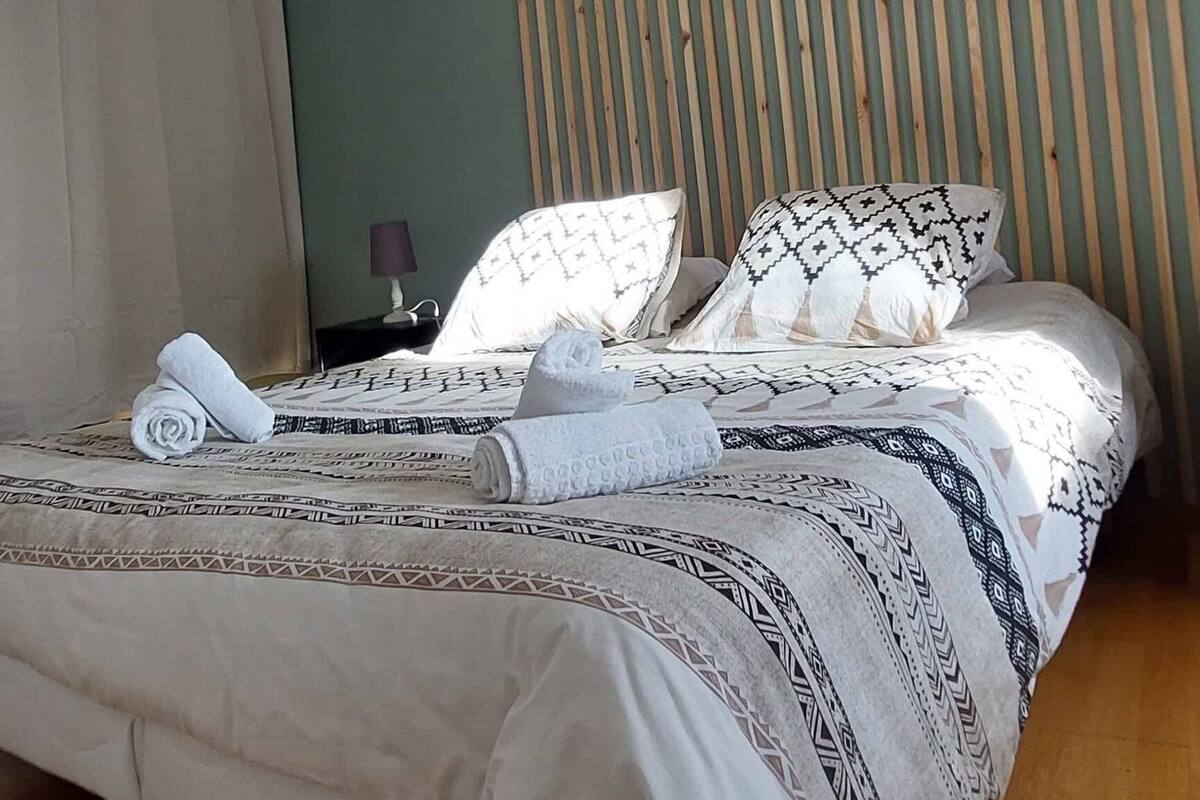 A modern bedroom with a slatted wooden ceiling, a bed with patterned bedding, and a nightstand with a lamp.