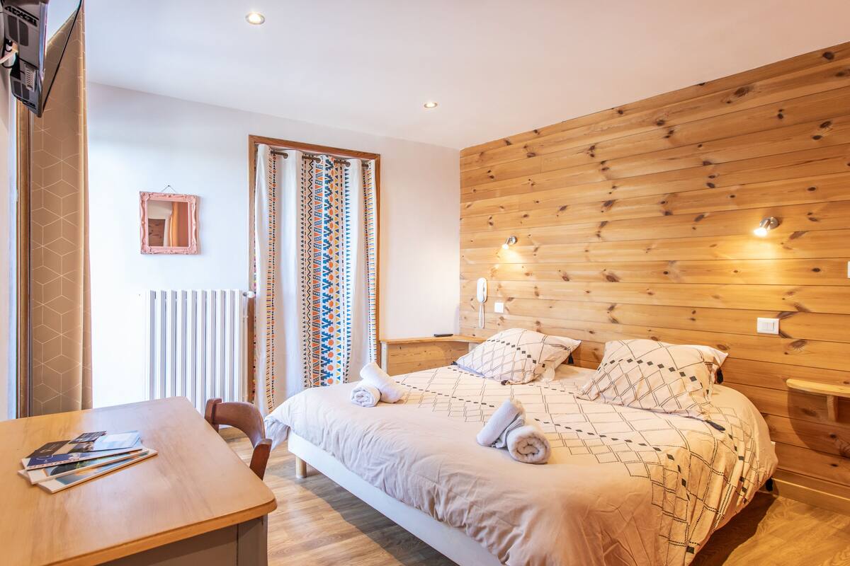 A neatly arranged bedroom with a wooden headboard, a bed with patterned bedding, a bedside table with a lamp, and a window with curtains.