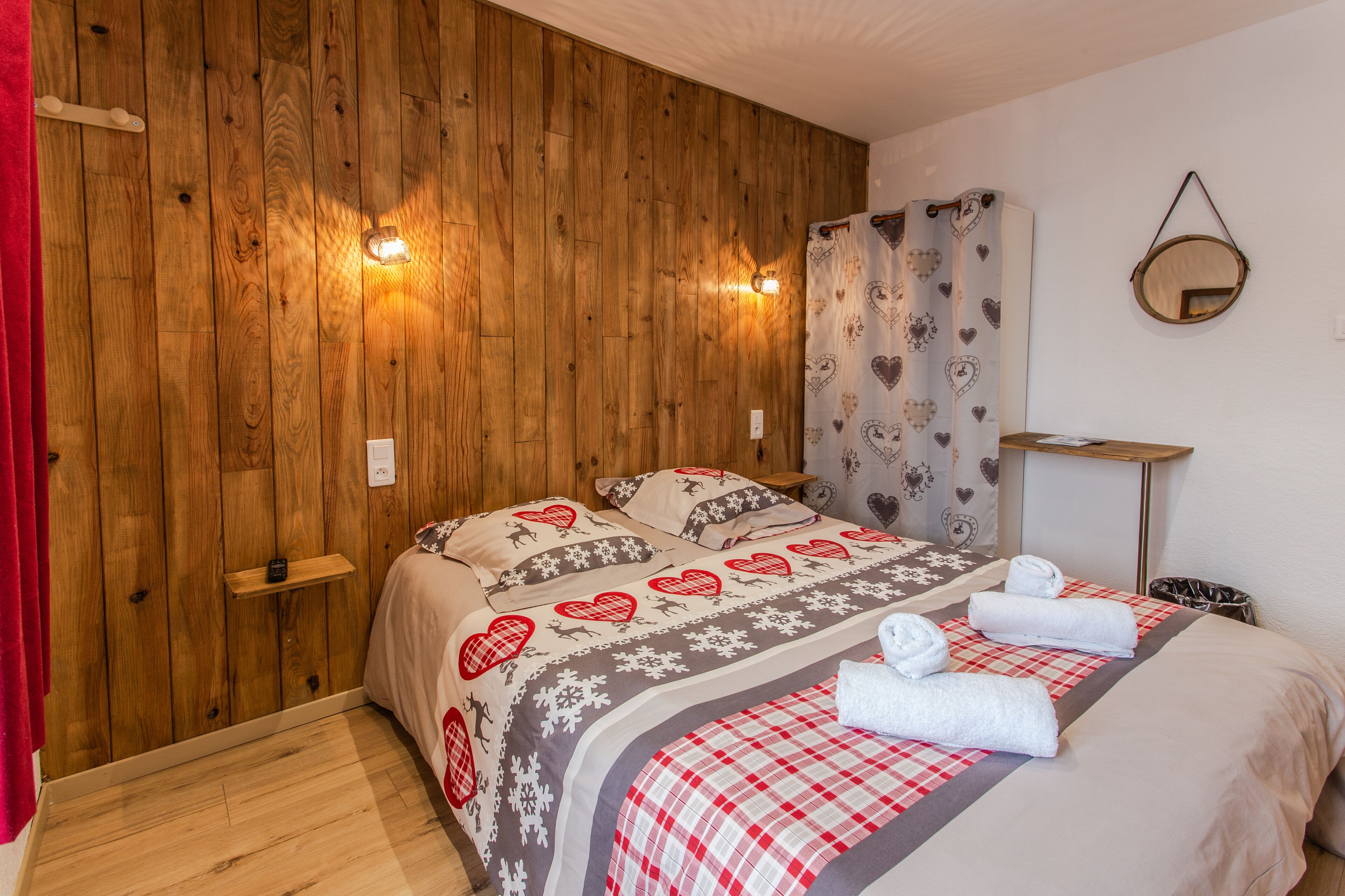 A bedroom with wooden panel walls, a bed with heart-patterned bedding, a bedside table, a mirror, and a red curtain.
