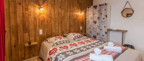 A bedroom with wooden panel walls, a bed with heart-patterned bedding, a bedside table, a mirror, and a red curtain.
