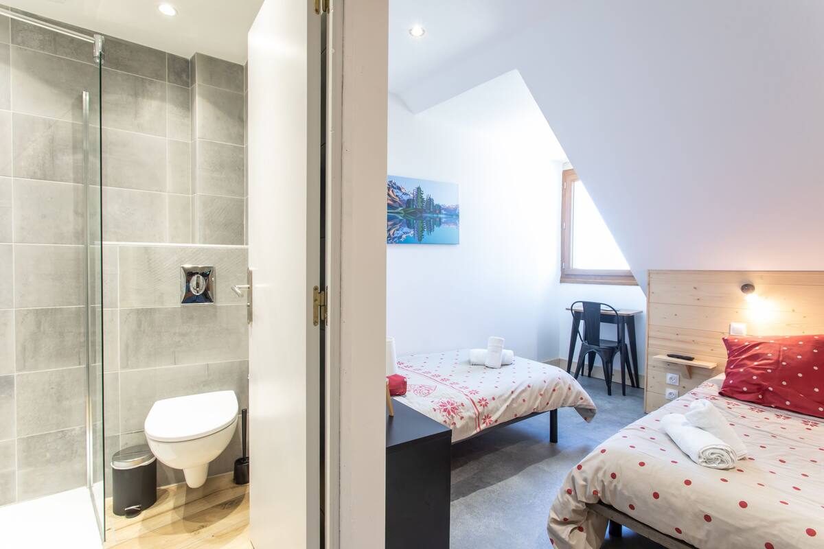 A modern hotel room with a shower, toilet, two beds with red polka dot bedding, a desk, and a painting on the wall.