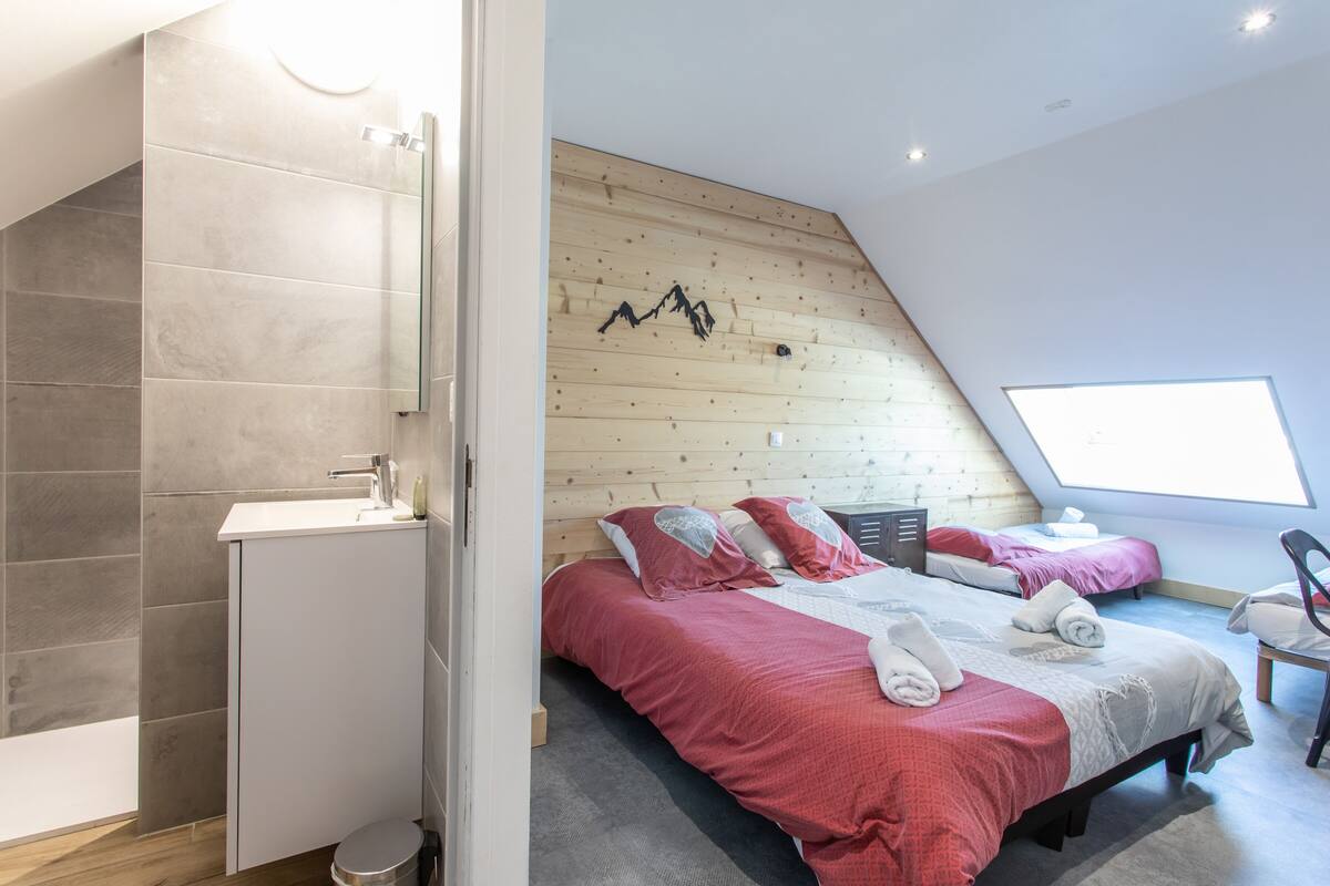 A modern bedroom with a sloped ceiling, a wooden wall with a mountain design, two beds with red bedding, a white sink, and a window providing natural light.