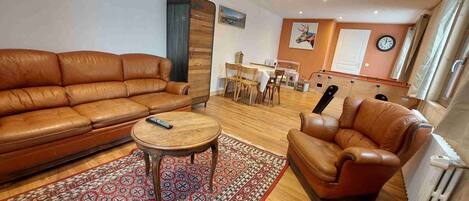 A living room with a brown leather sofa, a small wooden table, and a brown leather armchair. There is a wooden cabinet, a dining table with chairs, and a clock on the wall.
