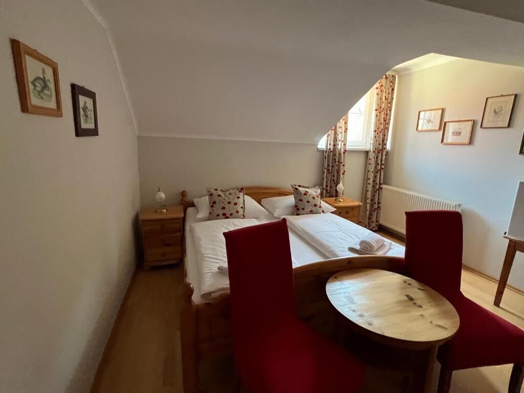 A bedroom with a bed, a nightstand, a red chair, a wooden table, and a window with curtains.