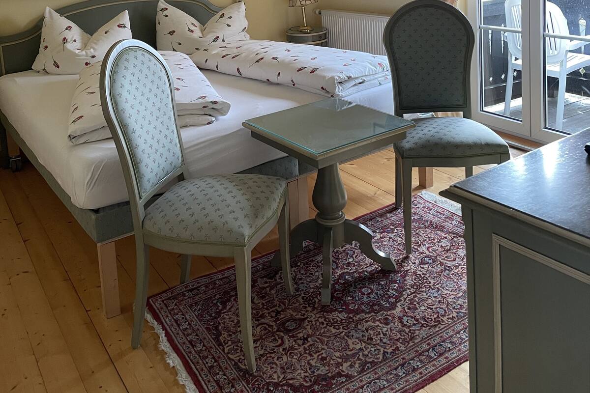Een slaapkamer met een bed, een stoel, een klein tafeltje, een radiator en een raam met gordijnen.