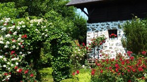 Un edificio de piedra con techo inclinado y chimenea, rodeado de vegetación exuberante y plantas con flores.