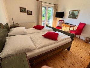 Een tweepersoonsbed met witte lakens en rode kussens, een houten vloer, een bureau met stoel, en een televisie aan de muur.