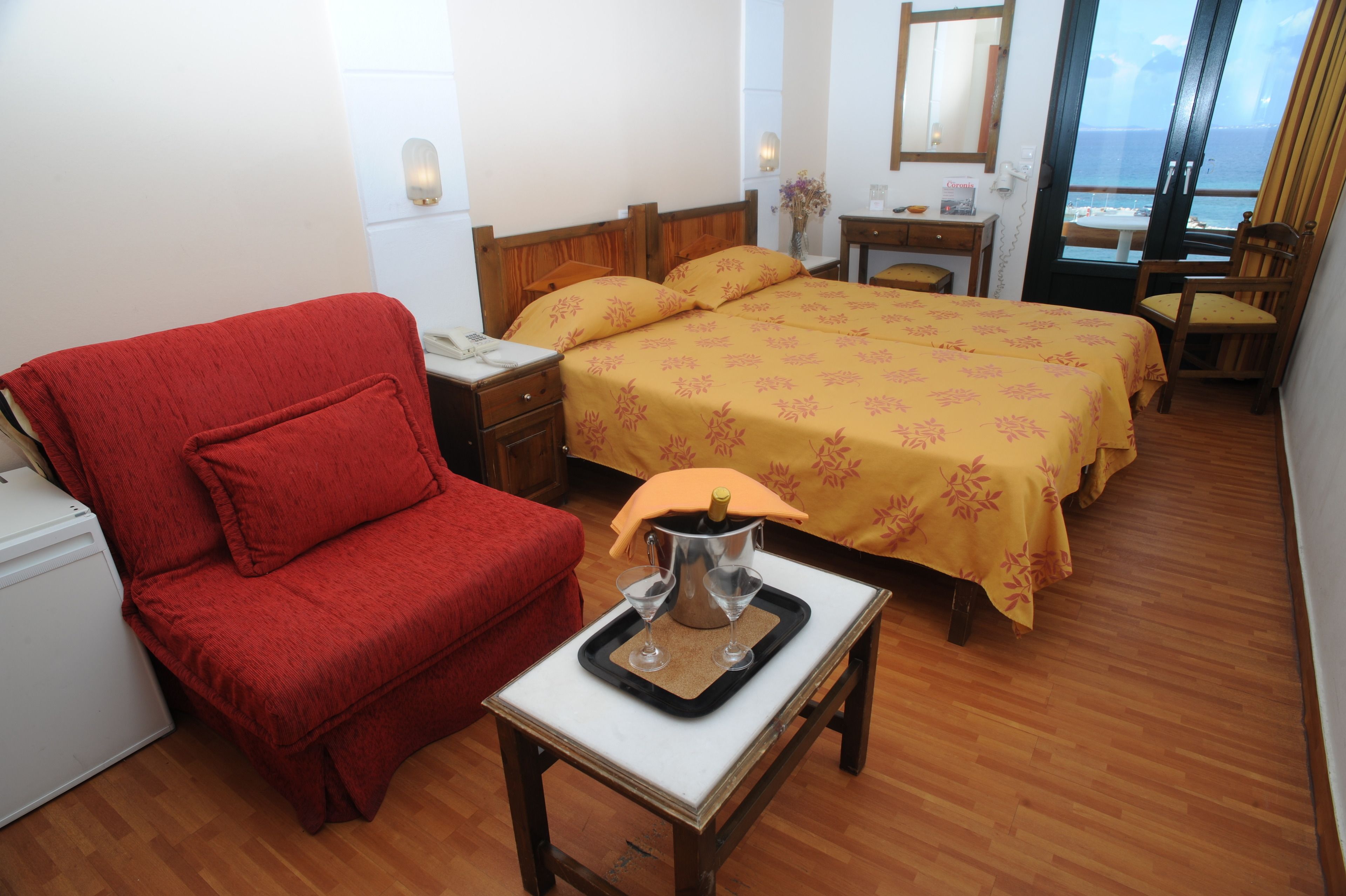 A hotel room with a wooden floor, a red armchair, two beds with yellow floral bedspreads, a small table with a tray holding a bottle and glasses, and a view of the sea through a large window.