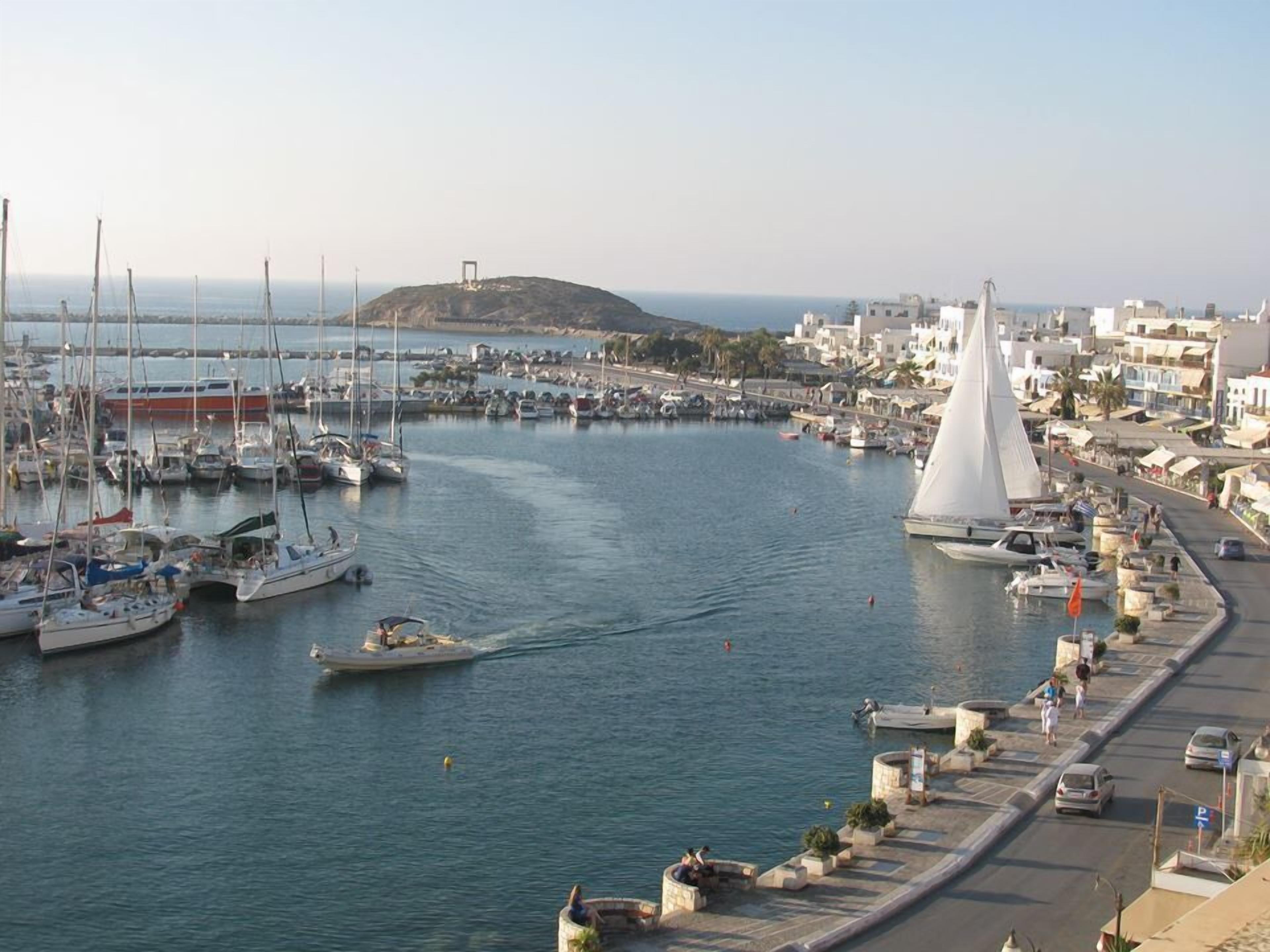 Un puerto deportivo con barcos amarrados, un velero con vela blanca, y un pueblo costero con edificios y una colina al fondo.