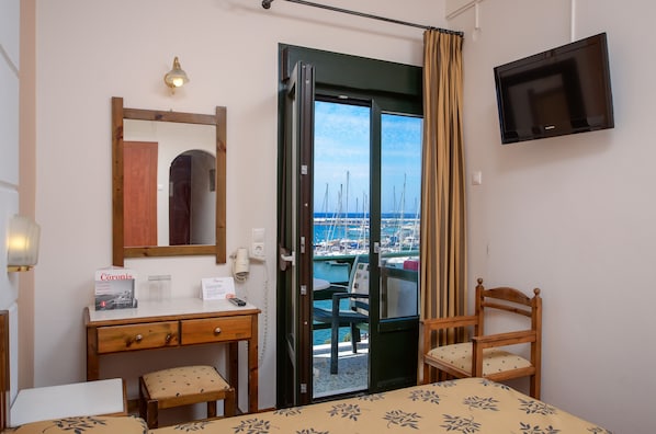 Habitación de hotel con cama, escritorio, silla, televisor, espejo y puerta que da a un balcón con vista al puerto deportivo.