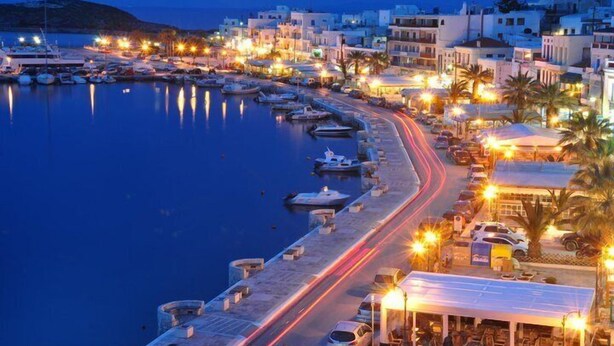 Un pueblo costero al anochecer, con un puerto deportivo, edificios a lo largo del paseo marítimo y una carretera con estelas de luz.