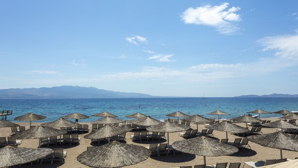 A beach with rows of sun loungers and thatched umbrellas, a clear sky, and a calm sea with distant mountains. - Charm Beach Hotel - All Inclusive (Bodrum)