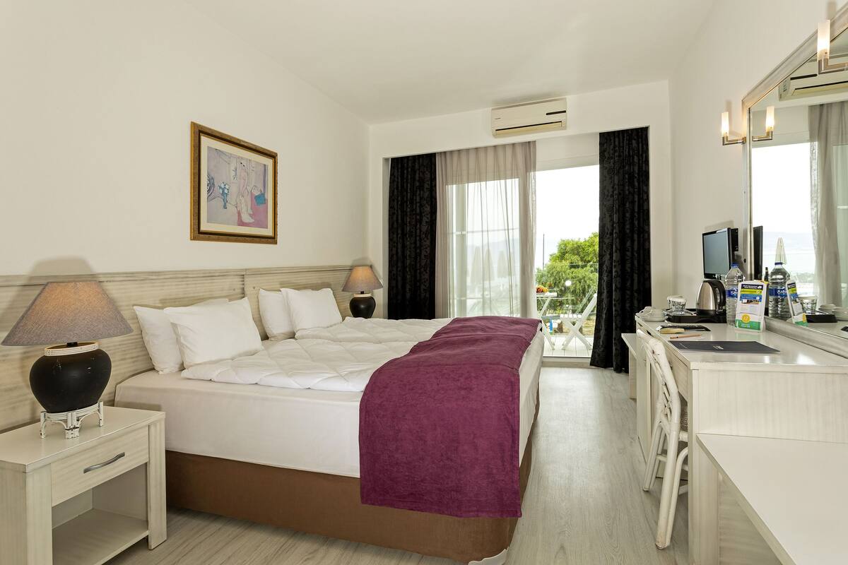 Quarto de hotel com cama, mesa de cabeceira, luminária, televisão e vista para o exterior.