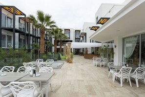 Un espace de restauration en plein air, avec des chaises et des tables en osier blanc, entouré de bâtiments modernes dotés de balustrades en verre et de palmiers.