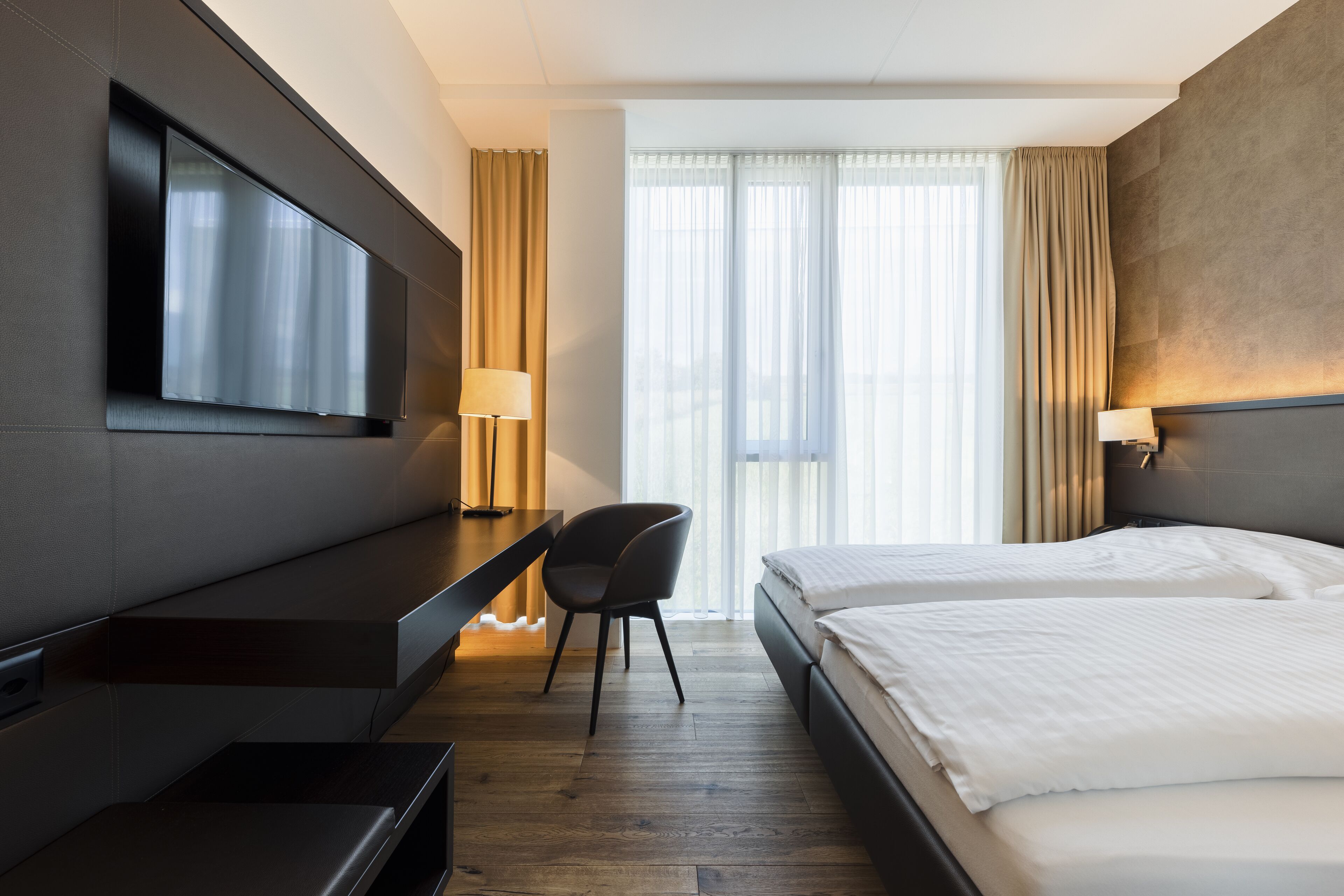 A hotel room with a bed, a desk, a chair, a TV, and a window with curtains.