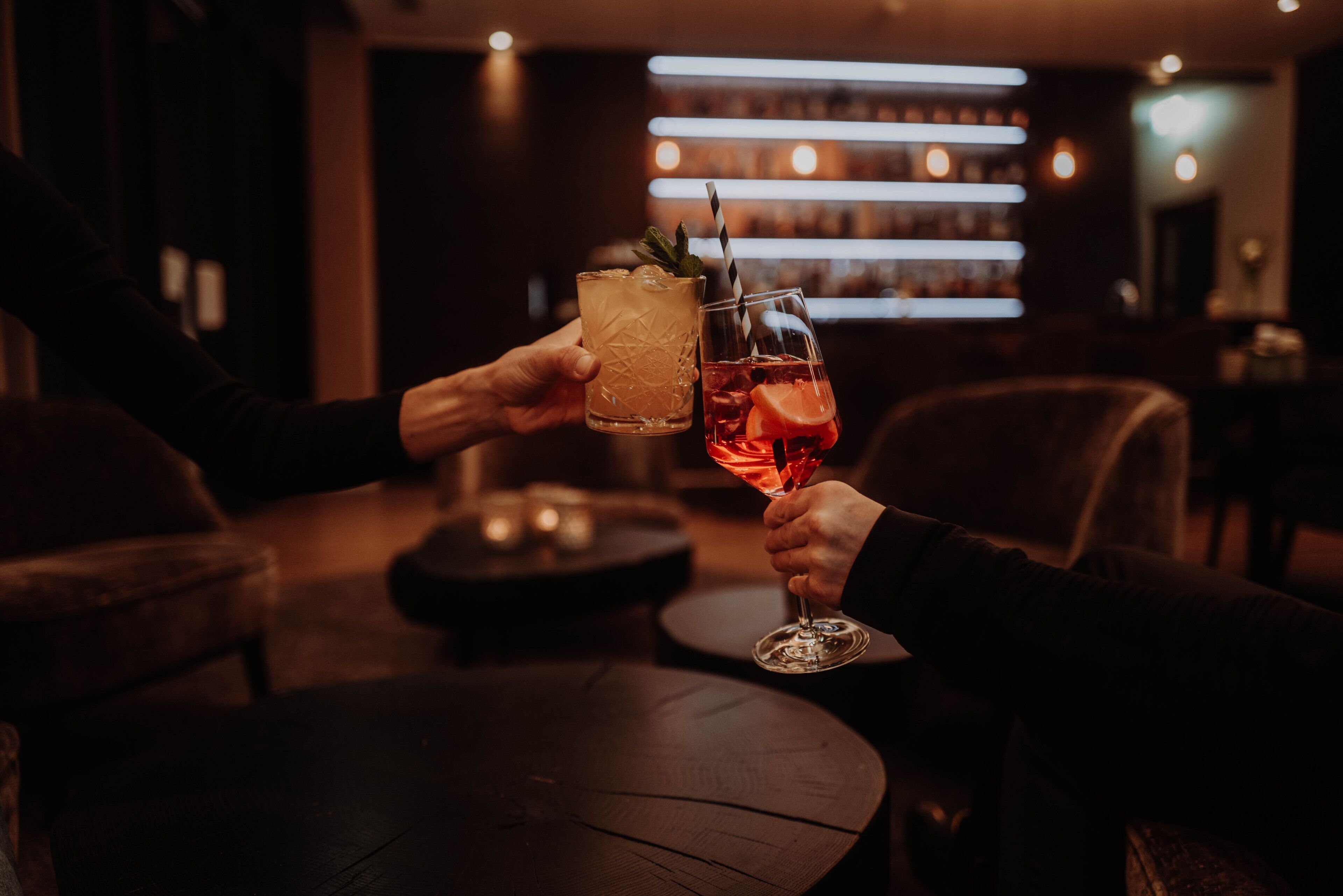 To personer holder glas med drikkevarer i en bar med mørk indretning og en hylde med flasker i baggrunden.