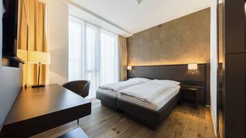 Habitación de hotel con cama, escritorio, silla y un ventanal con cortinas.