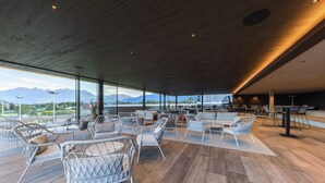 A spacious indoor area with modern wicker furniture, large windows offering mountain views, and a wooden floor. - Hotel Kommod (Ruggell)