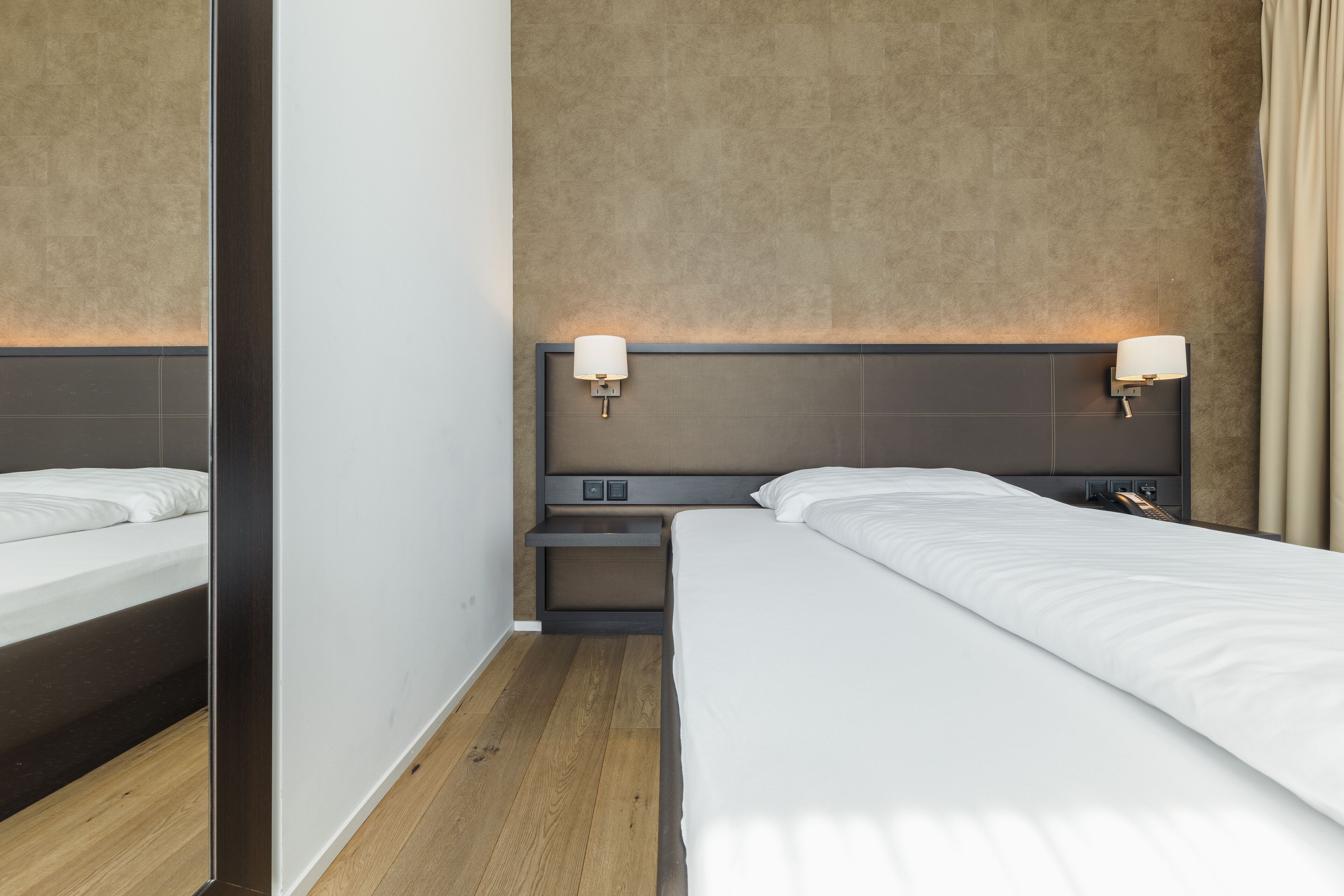 A modern hotel room with a large bed, a nightstand, a wall-mounted lamp, and a mirror.