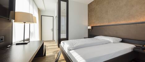 A modern hotel room with a wooden floor, a large bed with white linens, a desk with a lamp, and a flat-screen TV.