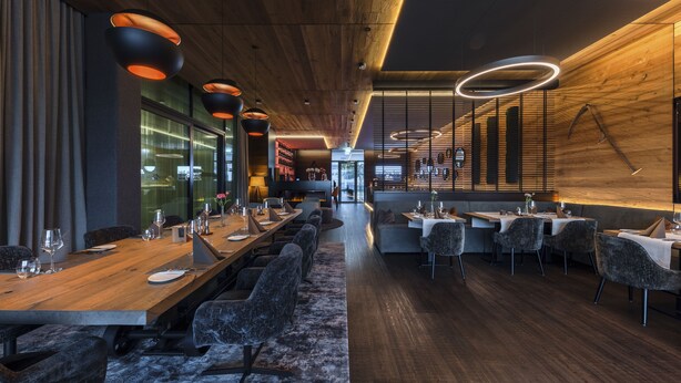 A modern restaurant with a long wooden table, multiple seating areas, and unique circular light fixtures. - Hotel Kommod (Ruggell)