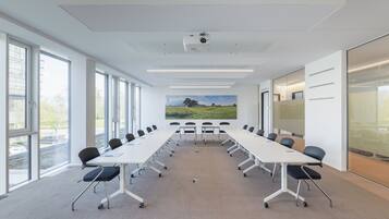 Uma sala de conferências com uma mesa comprida, cadeiras, um projetor e janelas amplas.