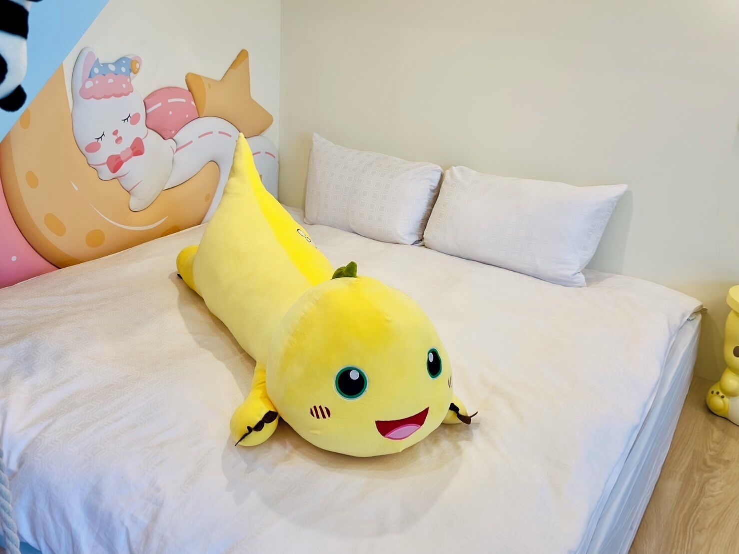 A bed with a yellow plush toy, white pillows, and a wall with a cartoon character.