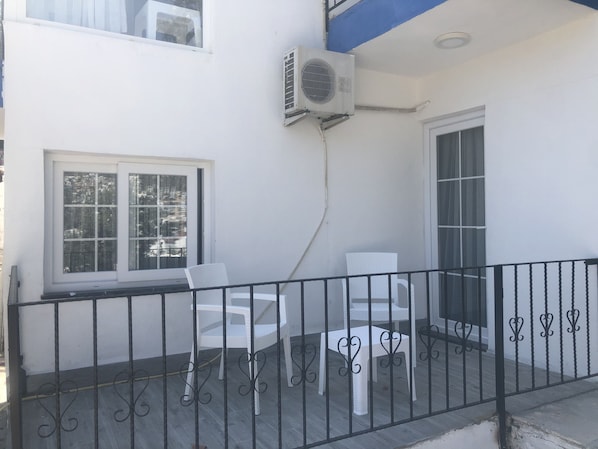 A balcony with white plastic chairs and a table, a window with a view of buildings, and an air conditioning unit mounted on the wall.