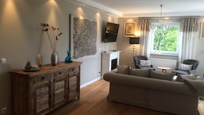 A living room with a sofa, wooden cabinet, TV, fireplace, and a window with curtains. - Luxus Ferienapartment “ Im Feldle „ (Kirchheim am Ries)