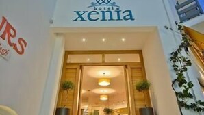 La entrada a un hotel llamado 'Xenia', con un frente de vidrio transparente, una escalera que lleva a la entrada, y un interior visible donde una persona está sentada a una mesa.