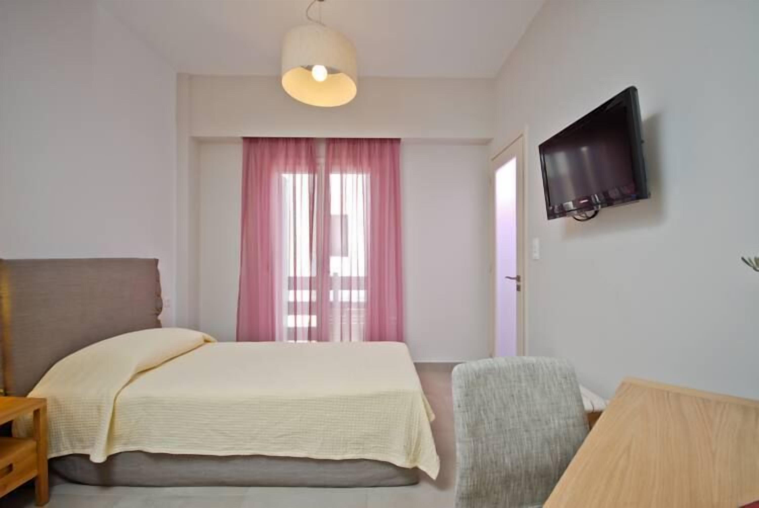 A bedroom with a bed, a chair, a television mounted on the wall, and a window with pink curtains.