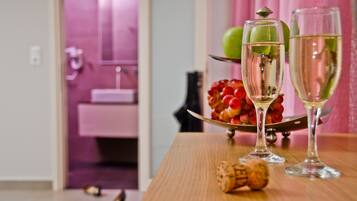 Ein Holztisch mit zwei Sektgläsern und einem Korkenzieher, eine Obstschale mit Äpfeln und Trauben sowie ein Badezimmer im Hintergrund.