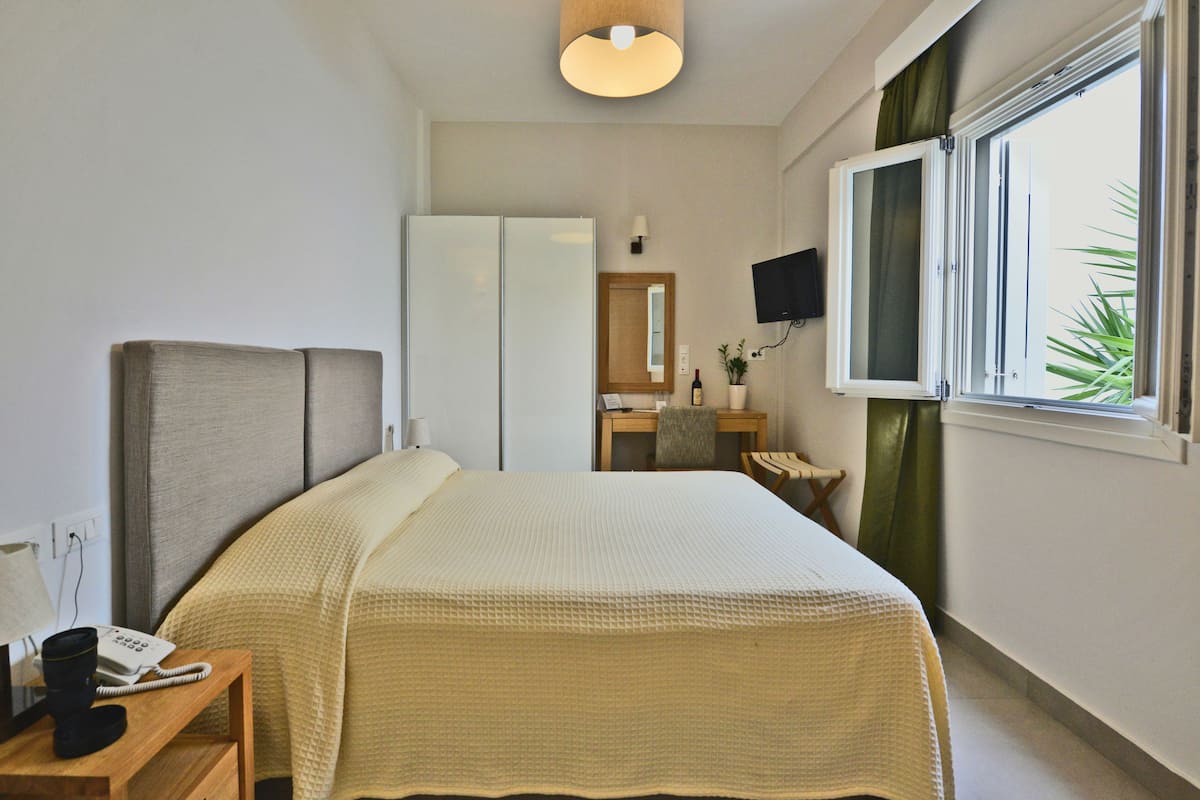 A hotel room with a bed, a desk with a telephone, a mirror, a television, and a window with curtains.