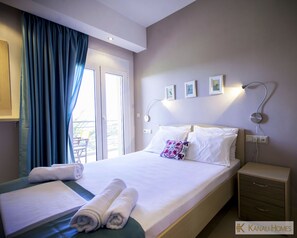 A hotel room with a bed, a nightstand, a desk, a chair, and a window with curtains. - Kanali Homes (Preveza)