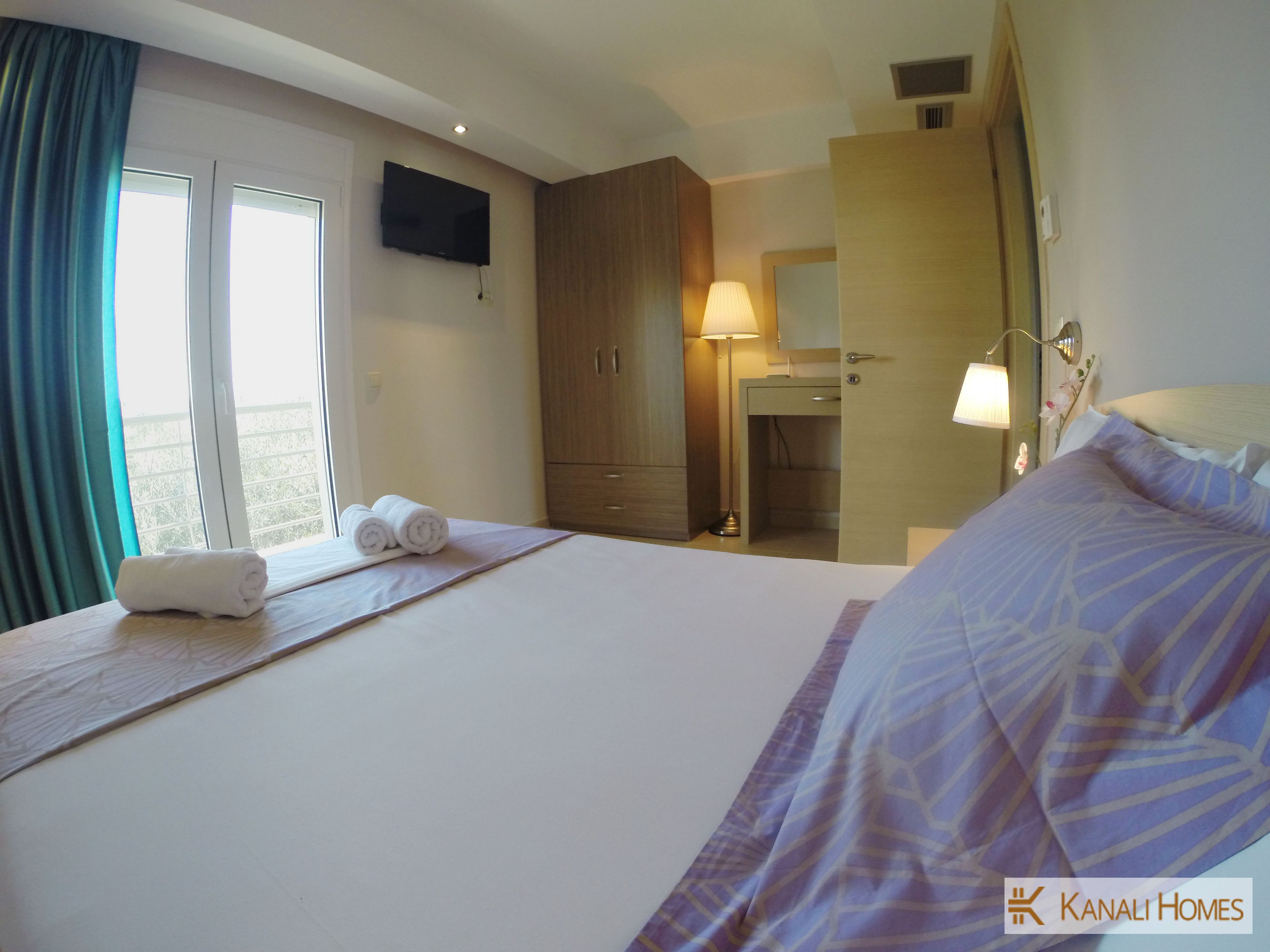A hotel room with a bed, a TV mounted on the wall, a wooden wardrobe, a bedside table with a lamp, and a window with curtains.