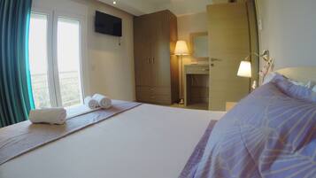 A hotel room with a bed, a TV mounted on the wall, a wooden wardrobe, a bedside table with a lamp, and a window with curtains.