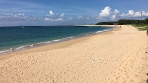 A wide sandy beach with gentle waves, a clear sky, and a distant shoreline with vegetation.