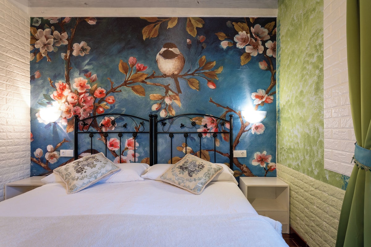 Ein Schlafzimmer mit floraler Wandmalerei, einem Bett mit weißer Bettwäsche, zwei Nachttischen und einem grünen Vorhang.