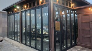 A glass-enclosed outdoor dining area with tables and chairs, surrounded by a wooden structure with a sloped roof and multiple lights. -  Aewol Oceanstay (Jeju City)