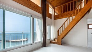 A spacious room with large windows overlooking a beach, a wooden staircase, and a white tiled floor. -  Aewol Oceanstay (Jeju City)