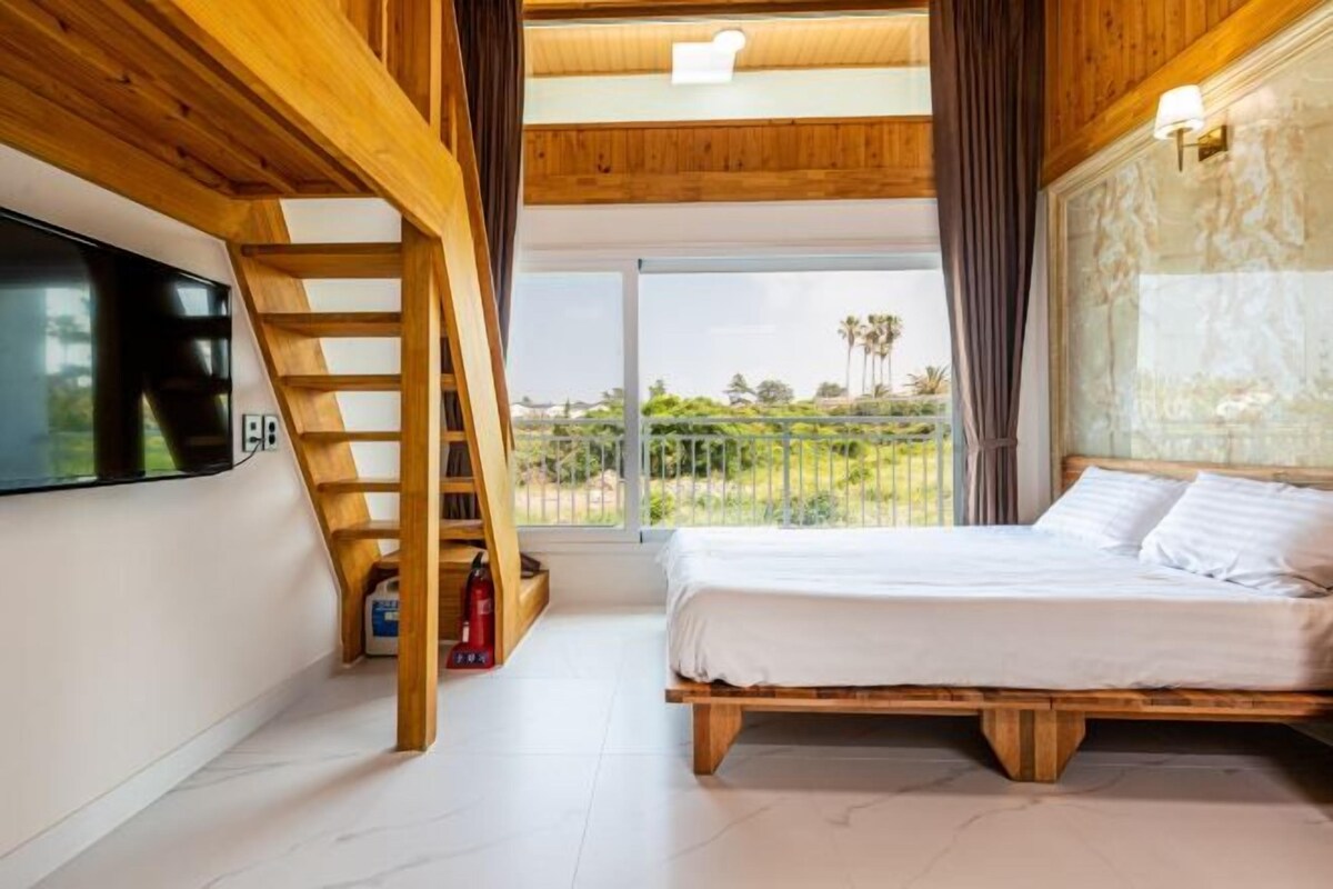 Une chambre avec un lit en bois, un téléviseur fixé au mur, une échelle en bois et un balcon donnant sur un paysage verdoyant.
