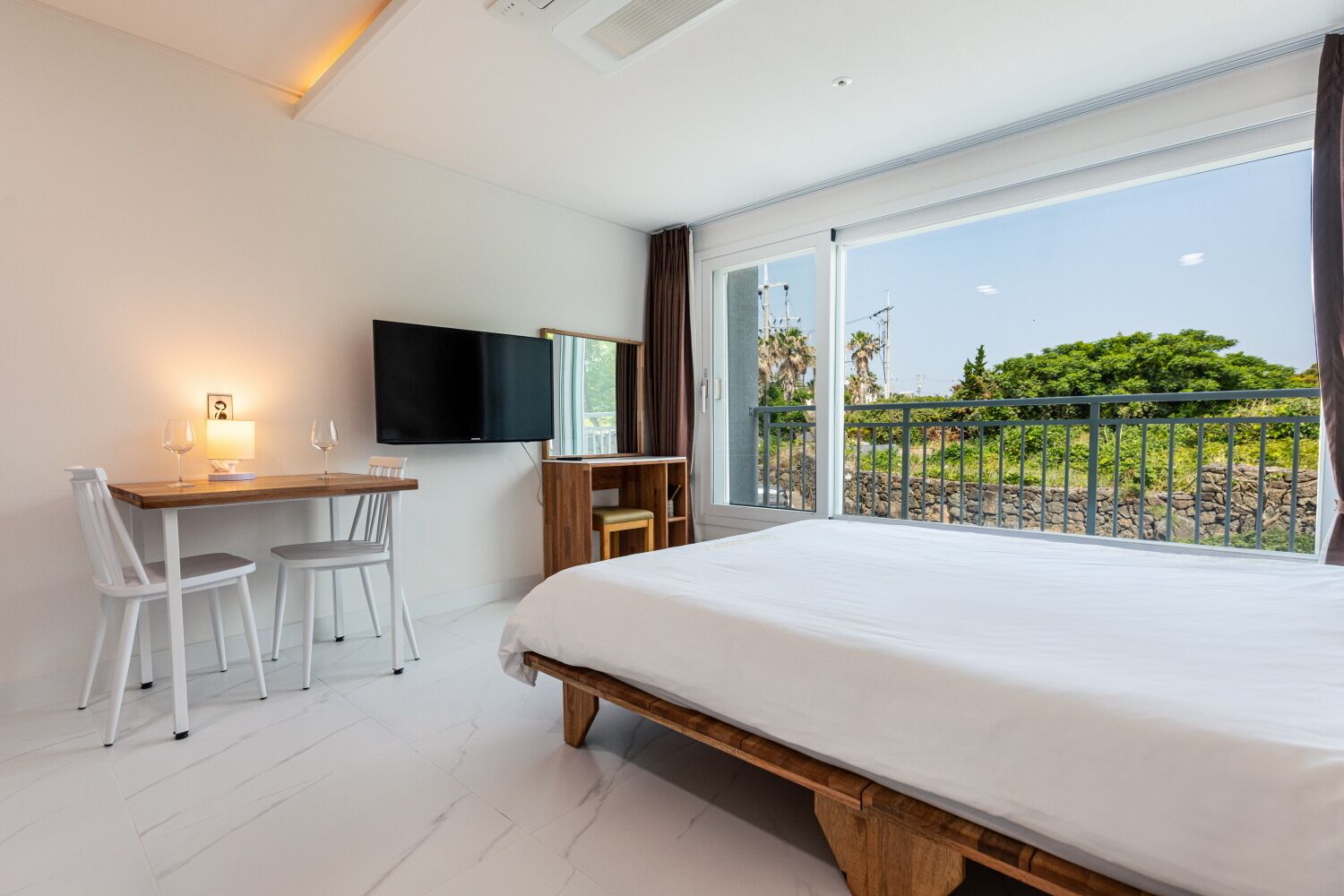 A modern hotel room with a large bed, a desk with two chairs, a flat-screen TV, and a balcony with a view of greenery.