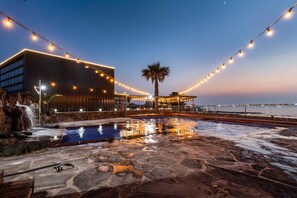 Un espace piscine agrémenté d’une cascade, de guirlandes lumineuses et d’un bâtiment moderne doté de grandes fenêtres.