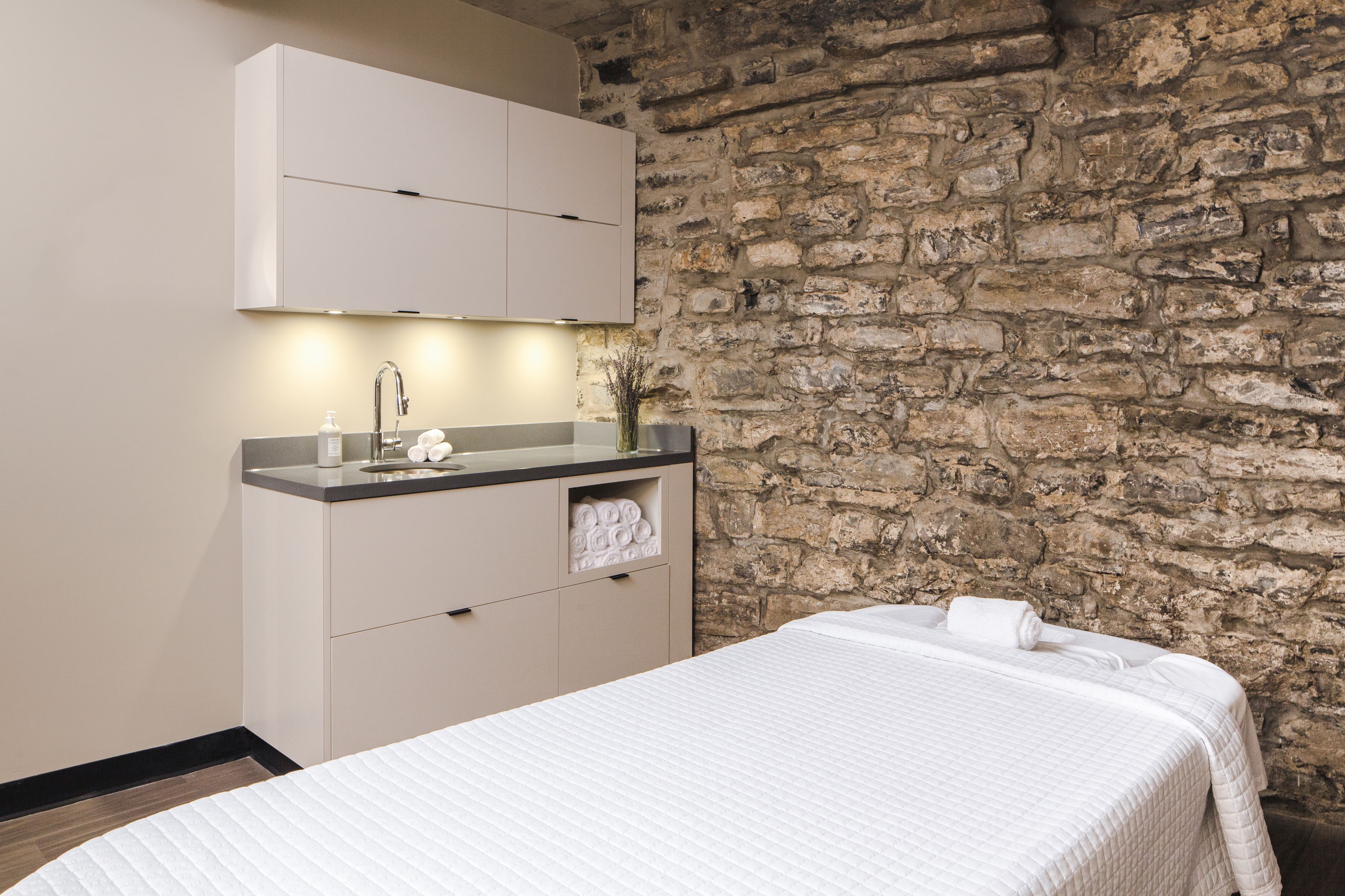 Un dormitorio con pared de piedra, una cama con edredón blanco, una mesita de noche, un espejo y un armario pequeño con artículos de tocador.