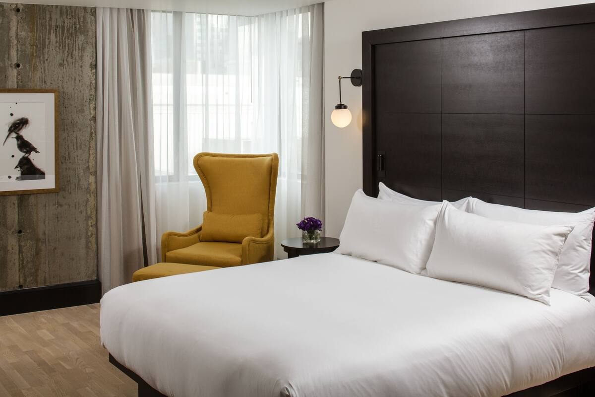 Une chambre d’hôtel avec un grand lit, un fauteuil de couleur moutarde, une petite table sur laquelle est posé un vase de fleurs et une tête de lit sombre.