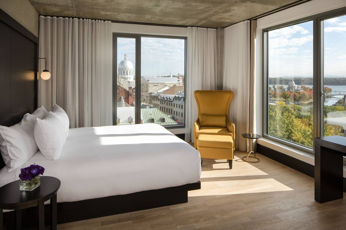 Une chambre d’hôtel avec un grand lit, un fauteuil jaune, une petite table sur laquelle est posé un vase de fleurs, et de grandes fenêtres donnant sur la ville.