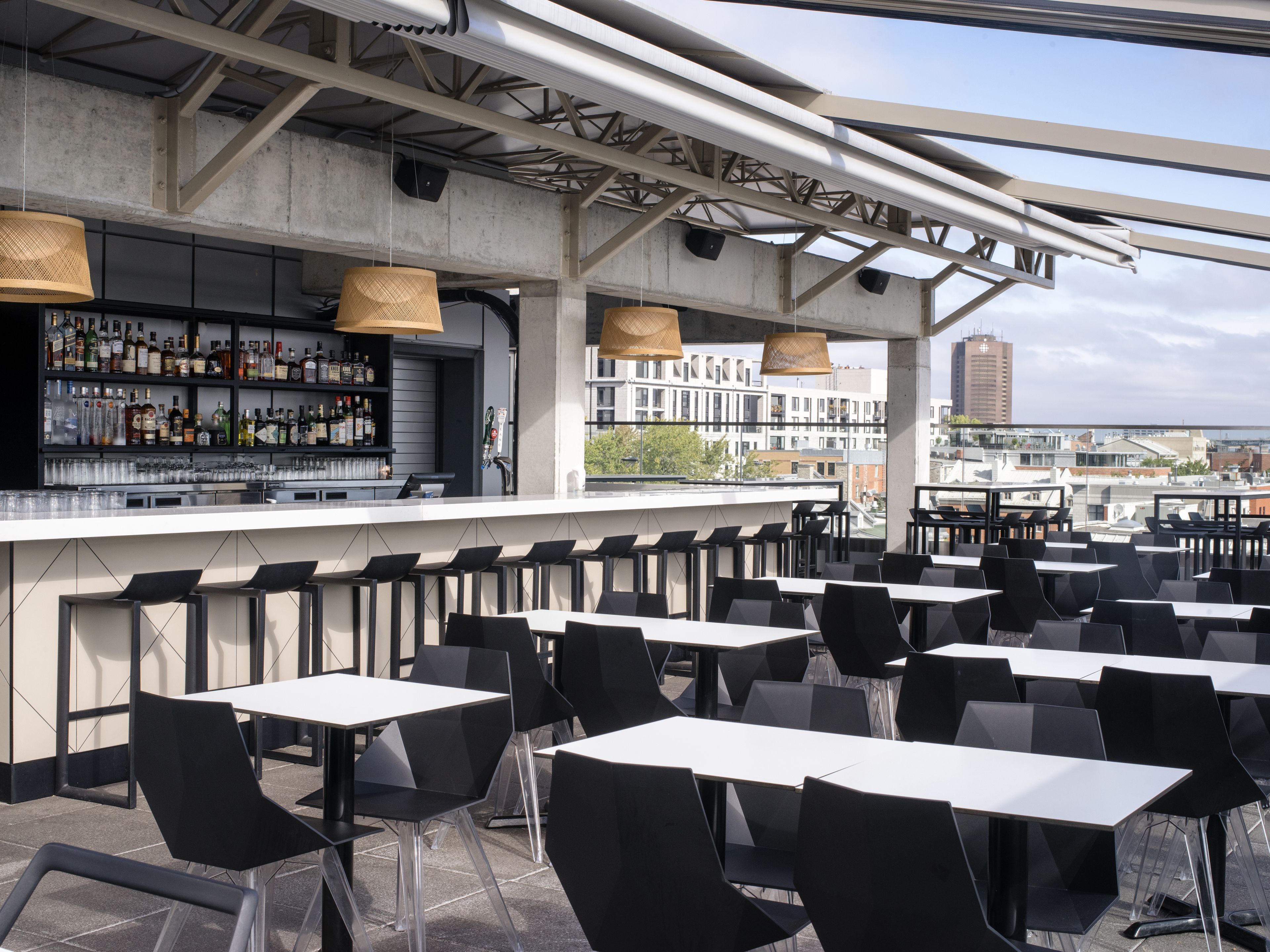 A rooftop bar with a bar counter, shelves of bottles, and black chairs and tables.