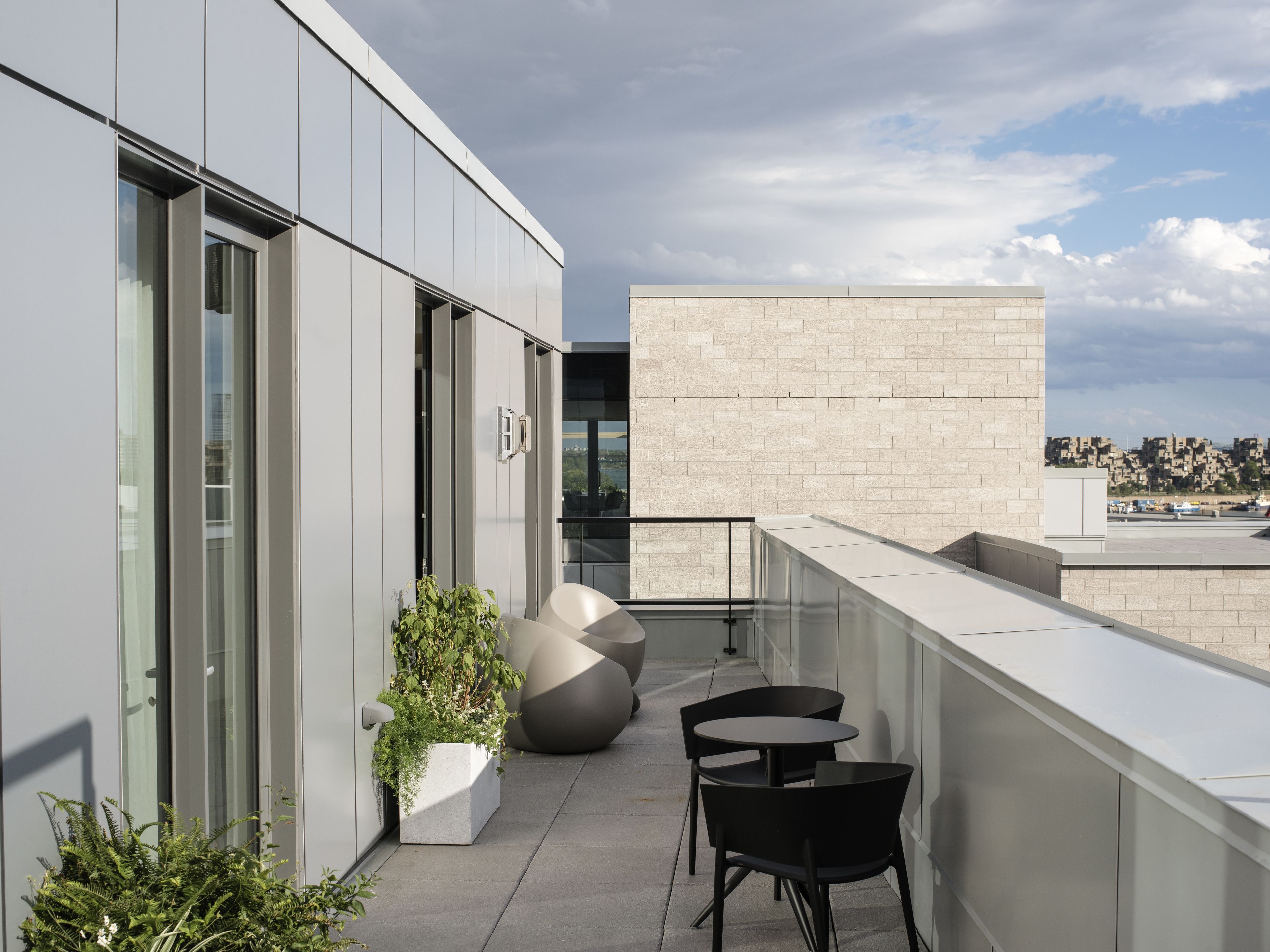 Une terrasse sur le toit, dotée d’un design moderne, comprenant un espace salon avec deux tables et des chaises, un grand bac à plantes agrémenté de verdure et offrant une vue sur le paysage urbain en arrière-plan.