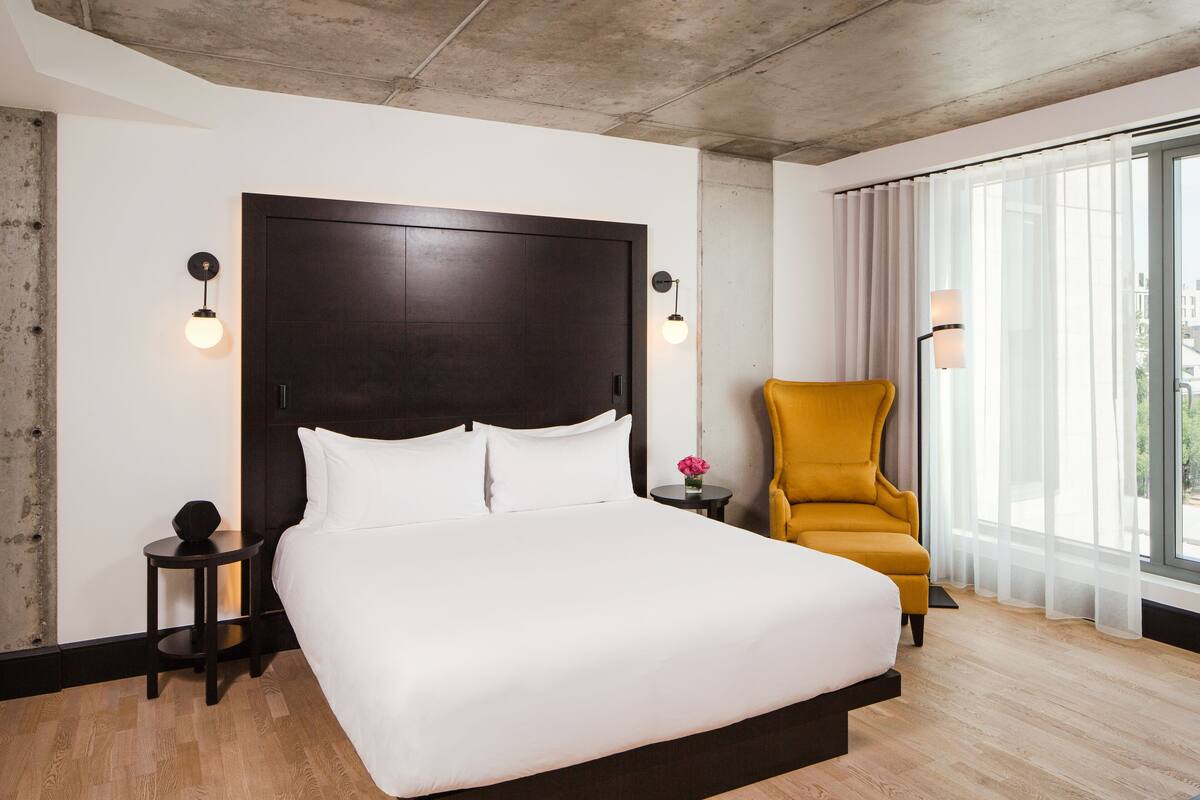 Une chambre d’hôtel moderne avec un grand lit, une tête de lit en bois, deux tables de chevet avec des lampes, un fauteuil jaune et une grande fenêtre avec des rideaux légers.