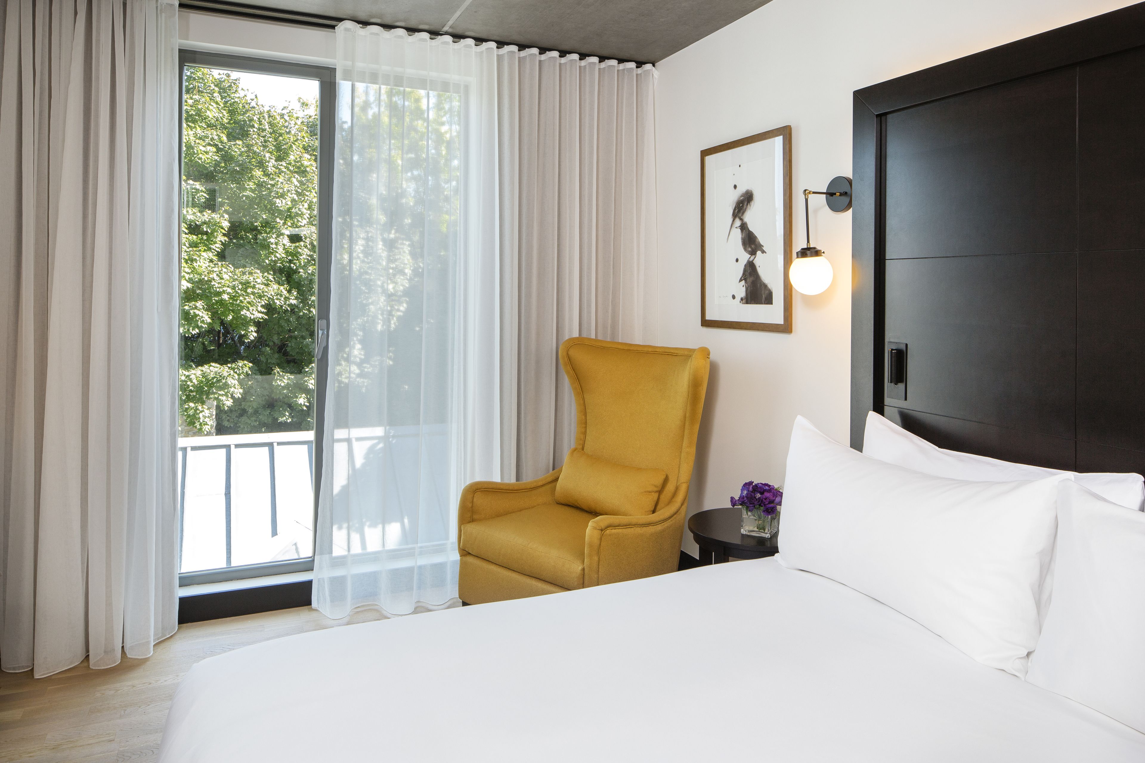 A hotel room with a bed, a yellow armchair, a small table with a vase of flowers, a framed picture on the wall, and a window with sheer curtains.